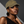 A woman wearing an olive green baseball cap with gold embroidered lettering that reads 'GOLD', paired with gold hoop earrings and a black one-shoulder top, set against a dark gray background with dramatic lighting.