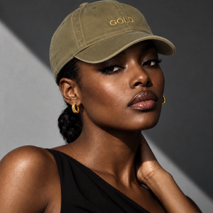 A woman wearing an olive green baseball cap with gold embroidered lettering that reads 'GOLD', paired with gold hoop earrings and a black one-shoulder top, set against a dark gray background with dramatic lighting.